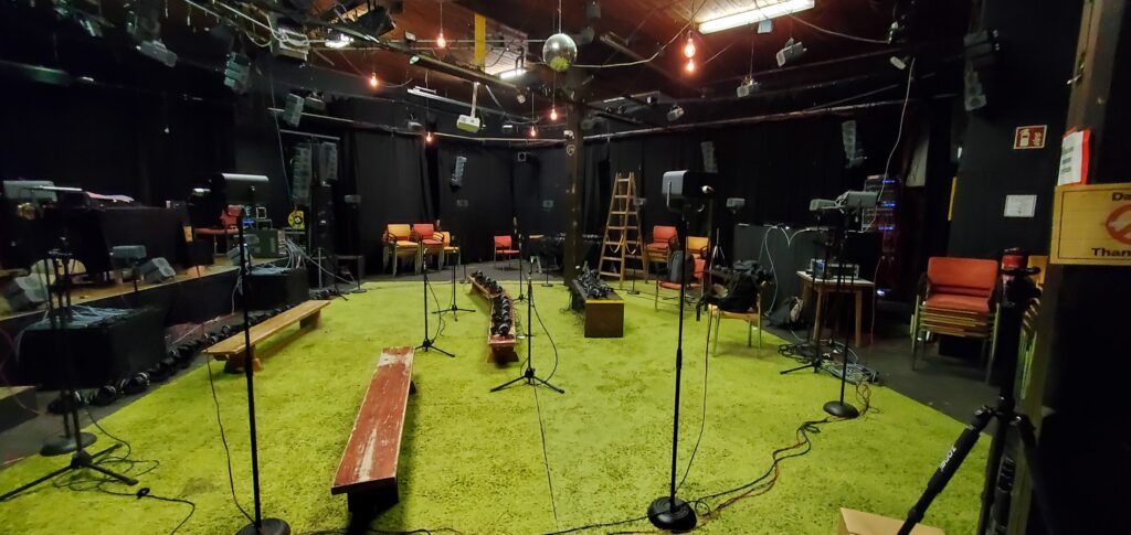 Hybrid audio diffusion setup with open headphones placed on a central bench and multiple loudspeakers arranged around the space before a performance.